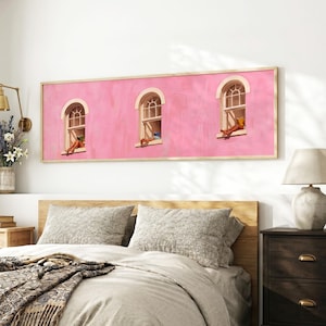 Puede incluir: Una pieza de arte mural rectangular que presenta una fachada rosa vibrante con tres ventanas arqueadas. Cada ventana muestra una pequeña figura descansando en el alféizar. La obra está enmarcada en madera clara y montada sobre una cama.