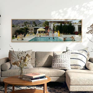 Puede incluir: Una sala de estar con una gran impresión enmarcada que representa una fiesta en la piscina de mediados de siglo. Un sofá seccional marrón claro con cojines estampados y una mesa de centro redonda de madera con libros están en primer plano. Una alfombra estampada completa la escena.