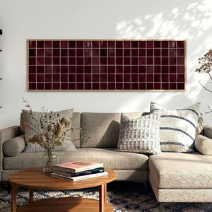 May include: A rectangular wall art piece featuring a grid of deep burgundy, glossy square tiles framed in light wood. The artwork is displayed horizontally above a beige sectional sofa, a wooden coffee table, and a textured area rug.