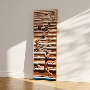 May include: A framed print depicting a vintage photograph of people sunbathing on tiered platforms. The image shows individuals in swimwear, with a warm, sunny colour palette. The artwork is displayed on a wooden floor against a white wall.