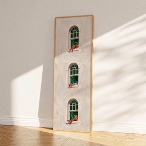 Puede incluir: Una obra de arte enmarcada verticalmente representa una fachada de edificio blanca con tres ventanas arqueadas. Cada ventana tiene cortinas verdes y una persona relajándose dentro. La obra está colocada contra una pared blanca y un suelo de madera.