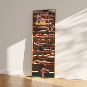 Puede incluir: Una impresión enmarcada que presenta una fotografía vintage de personas tomando el sol en escaleras. La imagen muestra a individuos en trajes de baño, con una paleta de colores cálidos. La obra de arte se muestra verticalmente, con un marco de madera.