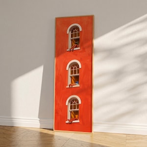 Op de afbeelding: Een verticale kunstdruk met een levendige oranje gebouwgevel met drie boogvensters. Elk venster toont een persoon die ontspant. Het kunstwerk is ingelijst in een dunne, lichtgekleurde houten lijst, leunend tegen een witte muur en een houten vloer.