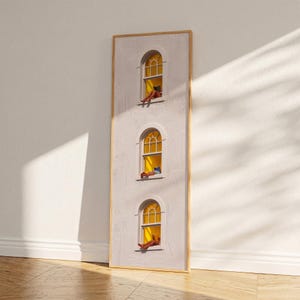 Puede incluir: Una impresión artística enmarcada representa una fachada blanca con tres ventanas arqueadas. Cada ventana muestra a una persona descansando, con luz amarilla en el interior. La obra está enmarcada en un borde dorado, apoyada contra una pared blanca.