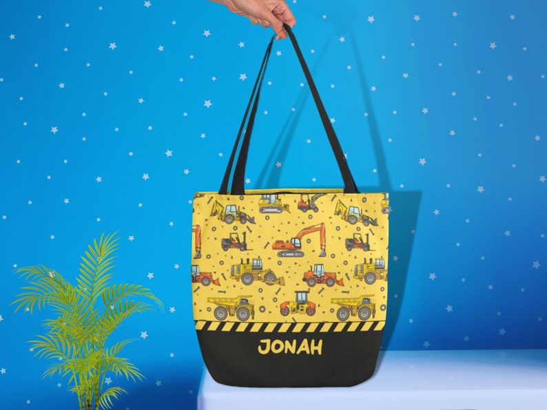May include: A yellow canvas tote bag with a black handle and a black base. The bag is decorated with a pattern of construction vehicles and the name "JONAH" is printed on the base in black.