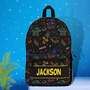 May include: Black backpack with a colorful space-themed pattern featuring rockets, planets, stars, and aliens. The backpack has a yellow name tag that says "JACKSON".