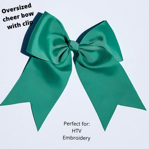 May include: An oversized teal cheer bow with a clip. The bow is made of a ribbed fabric and has long, pointed tails. Text on the image reads "Oversized cheer bow with clip."