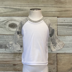 May include: White long-sleeve shirt with grey raglan sleeves and ruffle detailing on the sleeves. The shirt is on a mannequin.