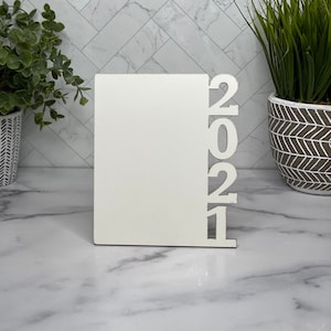 May include: White wooden picture frame with the year 2021 cut out on the right side. The frame is standing on a white marble surface.