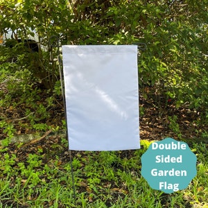 May include: A blank white double-sided garden flag. The flag is attached to a black metal pole and is displayed in a grassy area.