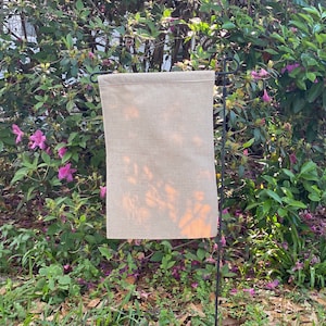 May include: A plain, beige burlap garden flag hanging on a black metal pole. The flag is blank and has a subtle texture. The flag is hanging in front of a green bush with pink flowers.
