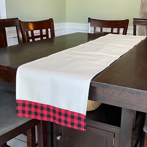 May include: A white table runner with a red and black buffalo plaid trim. The runner is on a dark wood table with two chairs on either side.
