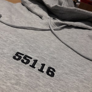 Op de afbeelding: Een grijs hoodie met het nummer 55116 geborduurd op de voorkant.