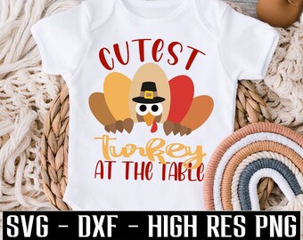 Puede incluir: Body de bebé blanco con un diseño de pavo colorido con un sombrero de peregrino. El texto en el body dice "Cutest Turkey at the Table". La parte inferior del body tiene el texto "SVG - DXF - HIGH RES PNG".