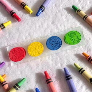 May include: A clear rectangular acrylic block featuring four circular embossed character portraits in red, yellow, blue, and green, surrounded by an assortment of colorful wax crayons scattered across a textured white surface.