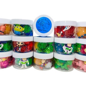 May include: Twelve clear plastic jars with white lids filled with colorful slime, beads, and small toys. The jars are arranged in three rows of four. The jars contain a variety of characters, including Buzz Lightyear, Woody, and other Disney characters. The jars are labeled "Thank You for Coming! Glitter Jars &"
