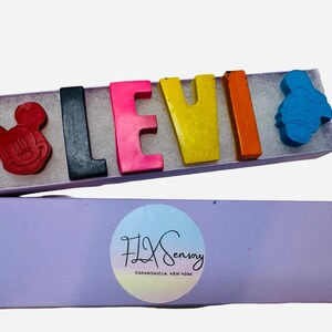 May include: A box of colorful crayons spelling the name "LEVI". The crayons are shaped like letters and cartoon characters, including Mickey Mouse and Donald Duck. The box is lavender and has a circular logo with the text "FLX Sensory CANANDAIGUA, NEW YORK".