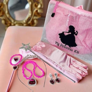 May include: A pink princess kit with accessories. The kit includes a mesh bag with a princess silhouette, a wand, a tiara, a necklace, earrings, and gloves. The accessories are pink and silver, with heart-shaped accents.