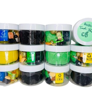 May include: Ten small clear jars with white lids filled with green, yellow, and black play dough. Each jar has small decorative items inside, such as gold glitter, miniature bottles, and plastic charms. The jars are stacked in three rows. The top jar has a green lid with the text "Bodhi's Trash Bash" in white.