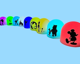 May include: A row of eight colorful light-up nightlights with different Disney character silhouettes. The characters include Tinkerbell, Mickey Mouse, Woody, Winnie the Pooh, and more.