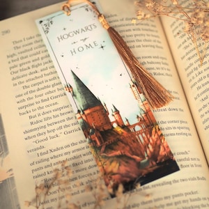 Puede incluir: Un marcap&aacute;ginas rectangular de papel con una ilustraci&oacute;n en acuarela de un castillo con torres altas. El texto "HOGWARTS is my HOME" est&aacute; impreso en la parte superior. Una borla dorada met&aacute;lica est&aacute; sujeta a la parte superior con una anilla, sobre un libro abierto.