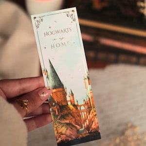 Peut inclure: Un marque-page rectangulaire en papier avec un fond de ciel bleu clair, des accents en feuille d'or et le texte HOGWARTS is my HOME. L'illustration représente un grand château avec de hautes tours et des ponts en pierre sur une falaise.