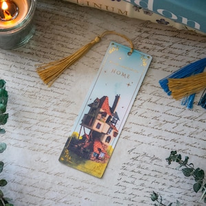 Peut inclure: Un marque-page doré avec un gland présente une illustration fantaisiste d'une maison avec un toit rouge et une porte bleue. Le marque-page a un fond doré avec le mot "HOME" imprimé en noir.