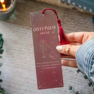 Peut inclure: Un marque-page bordeaux avec le texte "GRYFFINDOR HOUSE" et une illustration de lion. Le marque-page a un pompon rouge et la citation "Where dwell the brave at heart their daring, nerve, and chivalry, set Gryffindors apart."