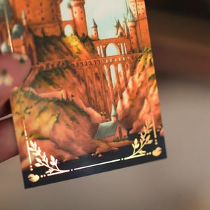 Puede incluir: Una tarjeta rectangular con una ilustraci&oacute;n detallada de un gran castillo con torres altas y un puente de piedra sobre un paisaje rocoso. La imagen est&aacute; enmarcada por un borde fino dorado con detalles de hojas en las esquinas inferiores.