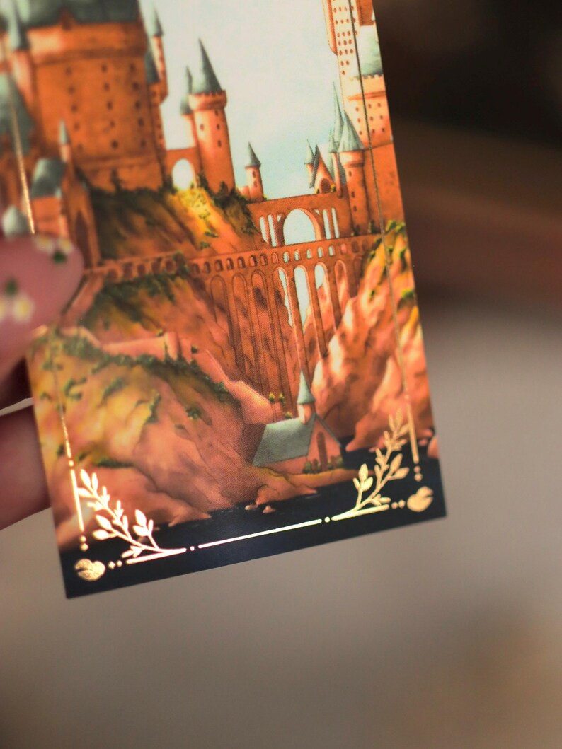 Puede incluir: Una tarjeta rectangular con una ilustraci&oacute;n detallada de un gran castillo con torres altas y un puente de piedra sobre un paisaje rocoso. La imagen est&aacute; enmarcada por un borde fino dorado con detalles de hojas en las esquinas inferiores.