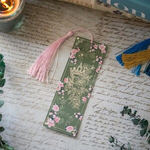 Puede incluir: Un marcap&aacute;ginas verde con una corona dorada y un dise&ntilde;o floral. El marcap&aacute;ginas tiene un borla rosa.