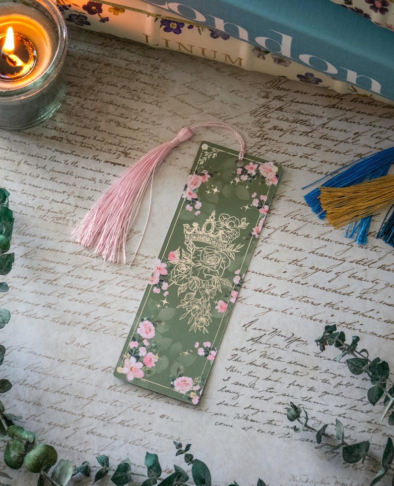 Puede incluir: Un marcap&aacute;ginas verde con una corona dorada y un dise&ntilde;o floral. El marcap&aacute;ginas tiene un borla rosa.