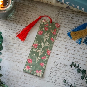 Puede incluir: Un marcap&aacute;ginas verde con un dise&ntilde;o floral y una flecha dorada. El marcap&aacute;ginas tiene un borla roja.
