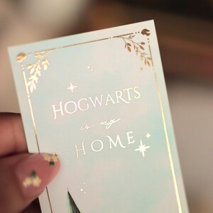 Puede incluir: Un marcap&aacute;ginas rectangular con fondo degradado azul claro y detalles en l&aacute;mina de oro. El texto HOGWARTS is my HOME aparece en letras doradas. El dise&ntilde;o incluye bordes de hojas doradas, peque&ntilde;as estrellas y una silueta de castillo verde.