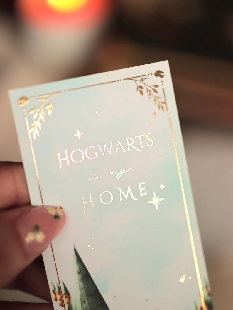 Puede incluir: Un marcap&aacute;ginas rectangular con fondo degradado azul claro y detalles en l&aacute;mina de oro. El texto HOGWARTS is my HOME aparece en letras doradas. El dise&ntilde;o incluye bordes de hojas doradas, peque&ntilde;as estrellas y una silueta de castillo verde.