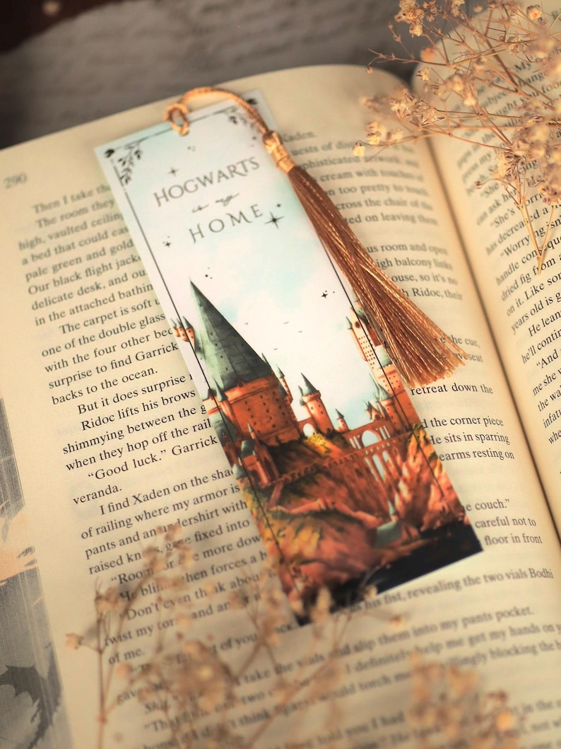 Puede incluir: Un marcap&aacute;ginas rectangular de papel con una ilustraci&oacute;n en acuarela de un castillo con torres altas. El texto "HOGWARTS is my HOME" est&aacute; impreso en la parte superior. Una borla dorada met&aacute;lica est&aacute; sujeta a la parte superior con una anilla, sobre un libro abierto.