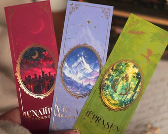 Puede incluir: Tres marcapáginas rectangulares con detalles en lámina de oro e ilustraciones ovaladas. El rojo muestra una ciudad, el azul montañas nevadas y el verde un bosque con astas. El texto incluye LUNATH, VELA y TERRASEN.