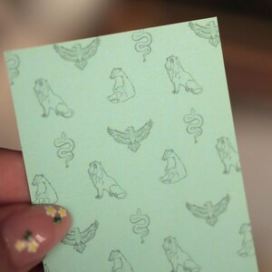 Puede incluir: Una tarjeta rectangular de color verde claro con un patr&oacute;n repetitivo de ilustraciones de l&iacute;neas verde oscuro. El dise&ntilde;o incluye representaciones de &aacute;guilas con las alas extendidas, serpientes enroscadas, leones sentados y tejones en cuadr&iacute;cula.