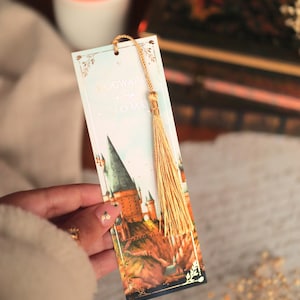 Puede incluir: Un marcap&aacute;ginas rectangular de papel con una ilustraci&oacute;n en acuarela de un castillo sobre un acantilado. El texto dice Hogwarts is my home. El marcap&aacute;ginas tiene un cord&oacute;n dorado y una borla dorada a juego en el orificio superior.