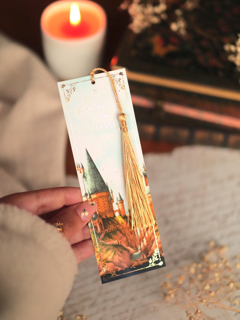 Puede incluir: Un marcap&aacute;ginas rectangular de papel con una ilustraci&oacute;n en acuarela de un castillo sobre un acantilado. El texto dice Hogwarts is my home. El marcap&aacute;ginas tiene un cord&oacute;n dorado y una borla dorada a juego en el orificio superior.