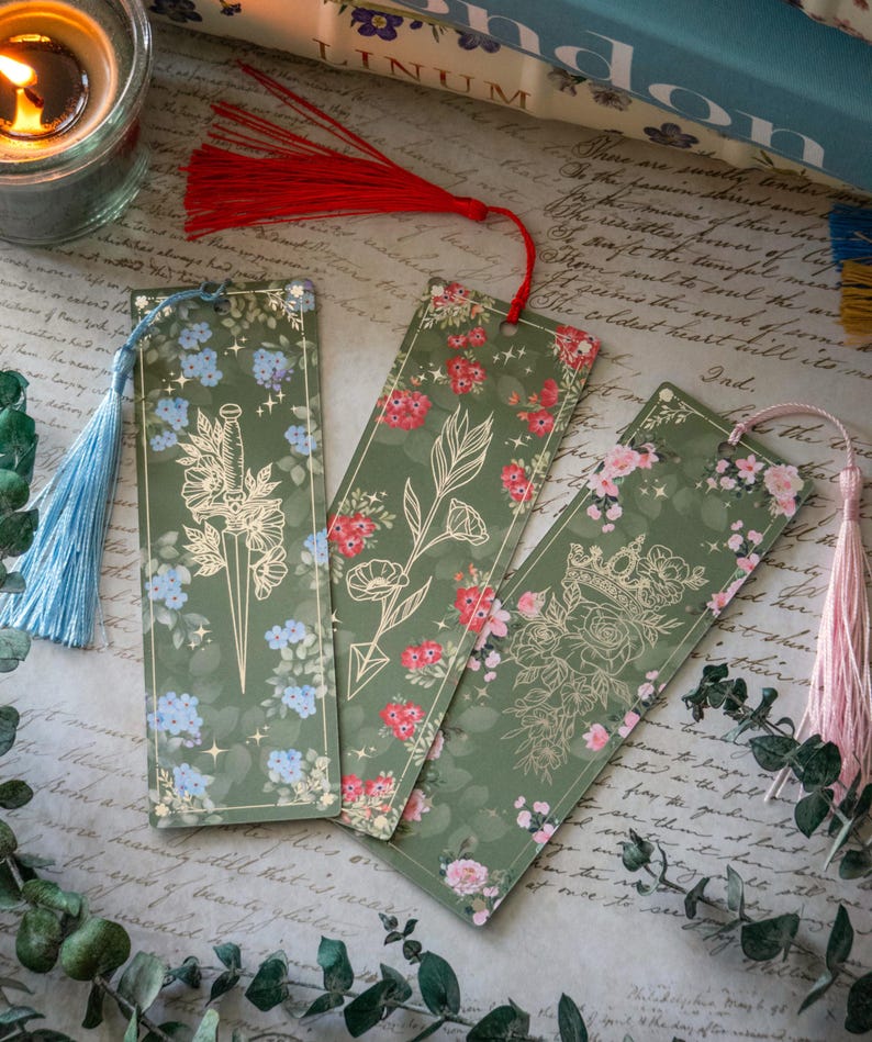 Puede incluir: Tres dise&ntilde;os de marcap&aacute;ginas verdes con ilustraciones florales doradas y borlas. Los dise&ntilde;os presentan una daga, una flecha y una corona.