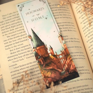 Puede incluir: Un marcap&aacute;ginas rectangular de papel con una ilustraci&oacute;n en acuarela de un castillo con torres altas y un puente. La parte superior tiene un fondo azul claro con el texto "HOGWARTS is my HOME" en negro, acentuado por peque&ntilde;as estrellas y bordes florales.
