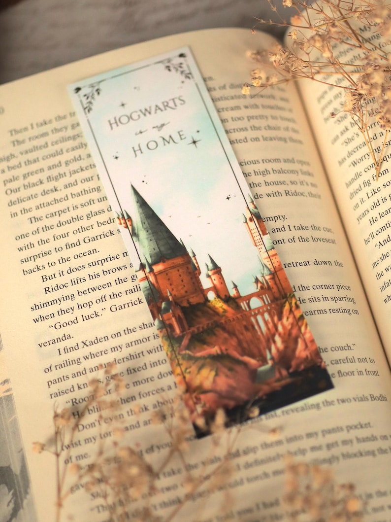 Puede incluir: Un marcap&aacute;ginas rectangular de papel con una ilustraci&oacute;n en acuarela de un castillo con torres altas y un puente. La parte superior tiene un fondo azul claro con el texto "HOGWARTS is my HOME" en negro, acentuado por peque&ntilde;as estrellas y bordes florales.