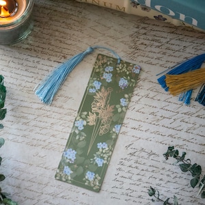 Puede incluir: Un marcap&aacute;ginas verde con un dise&ntilde;o de l&aacute;mina dorada de una daga y flores. El marcap&aacute;ginas tiene un borla azul.