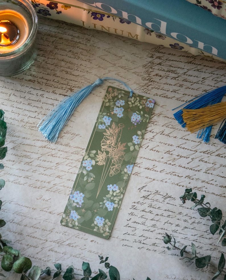 Puede incluir: Un marcap&aacute;ginas verde con un dise&ntilde;o de l&aacute;mina dorada de una daga y flores. El marcap&aacute;ginas tiene un borla azul.