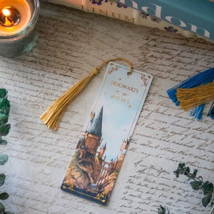 以下が含まれることがあります： ホグワーツ城の詳細なイラストと「HOGWARTS is HOME」の文字が描かれた装飾的なブックマーク。 金色のタッセルが付いており、手書きのスクリプトとろうそくの背景に配置されています。