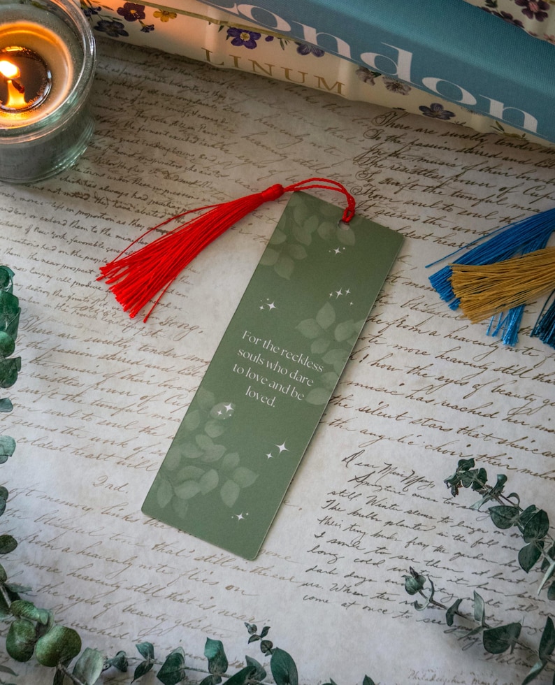 Puede incluir: Un marcap&aacute;ginas verde con un dise&ntilde;o de hojas y estrellas doradas. El marcap&aacute;ginas tiene un borla roja y el texto "Para las almas imprudentes que se atreven a amar y ser amadas."