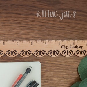 Op de afbeelding: Een houten liniaal met een decoratieve gegolfde rand en de tekst "Miss Lindsey" erop gegraveerd. De liniaal ligt op een houten oppervlak met een wit notitieboekje en pennen.