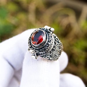 May include: A silver ring with an oval, deep red gemstone at the center. The ring features intricate, swirling filigree designs around the gemstone and along the band. The ring is held up by a white glove.