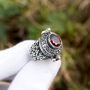 May include: A detailed silver ring with an oval, deep red gemstone. The ring features intricate scrollwork and floral patterns. The gemstone is set in a decorative bezel, and the ring appears to open. A unique piece of jewelry.